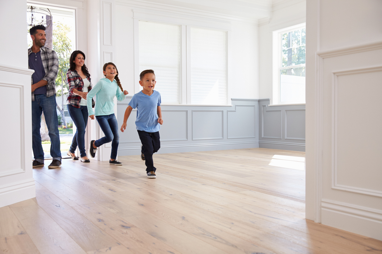 A joyful family of four—parents and two young children—entering a bright, modern home for the first time. The children excitedly run across the hardwood floors while the parents smile from the doorway, capturing the excitement of buying a new home.